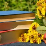 Stack of three books on a table outside next to a pot of yellow flowers and an apple