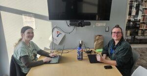 Our library social workers sit at their laptop computers on each side of a long table. They both have green shirts and there is a black TV monitor on the wall.