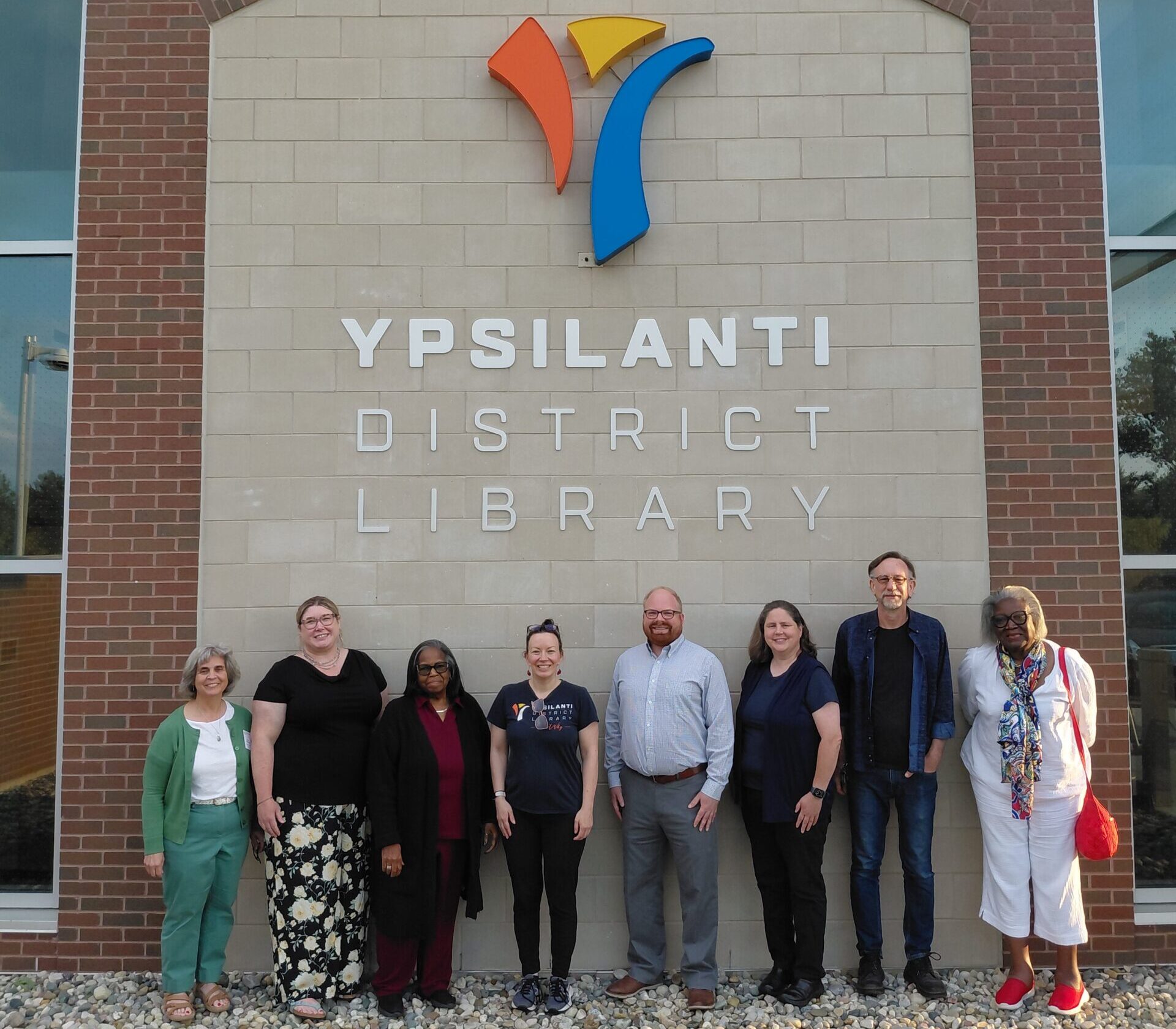 8 people posed in front of a brick wall with the Ypsilanti District Library name and logo on it.